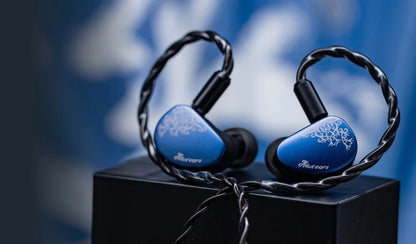 Blue and black in-ear earphones with decorative white floral patterns on the ear tips, resting on a black display block against a blurred blue background.