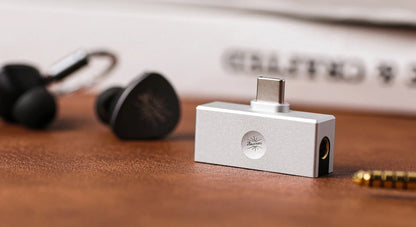 Allergo Mini DAC with a circular logo on the front, displayed on a wooden surface next to black in-ear earphones.