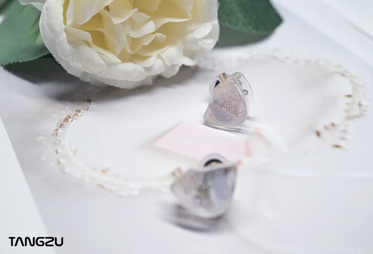 Pair of white IEMs displayed on a white surface with a white rose and delicate lace in the background. The brand name 'TANGZU' is visible in the bottom-left corner.
