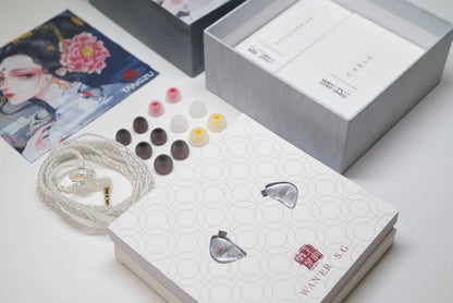 A pair of in-ear monitors with a white braided cable, shown alongside multiple ear tips, an open presentation box, and a branded package on a clean white surface.
