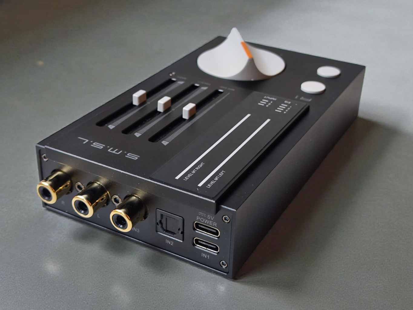 A Black SMSL R1 device with gold and silver connectors, sliding controls, and a white cone-shaped component on top, photographed on a gray surface.